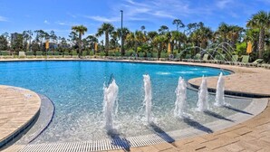 Piscina azul cristalina con múltiples chorros de agua, rodeada de palmeras y sillas de descanso.