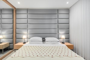 A bedroom with a tufted headboard, a bed with white linens, two bedside tables with lamps, and a large mirror on one wall.