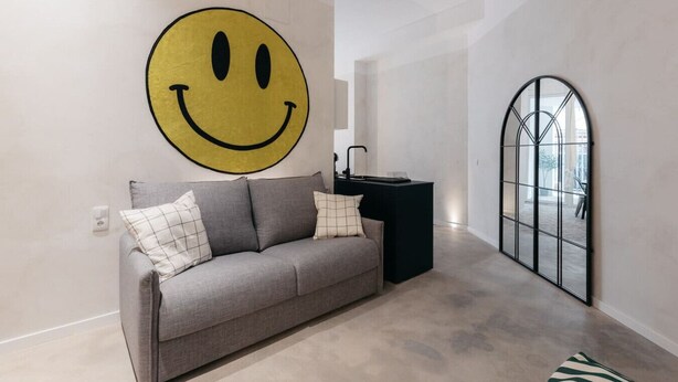 A modern living room with a large smiley face mural, a grey sofa with patterned cushions, a black side table, and a large arched mirror.