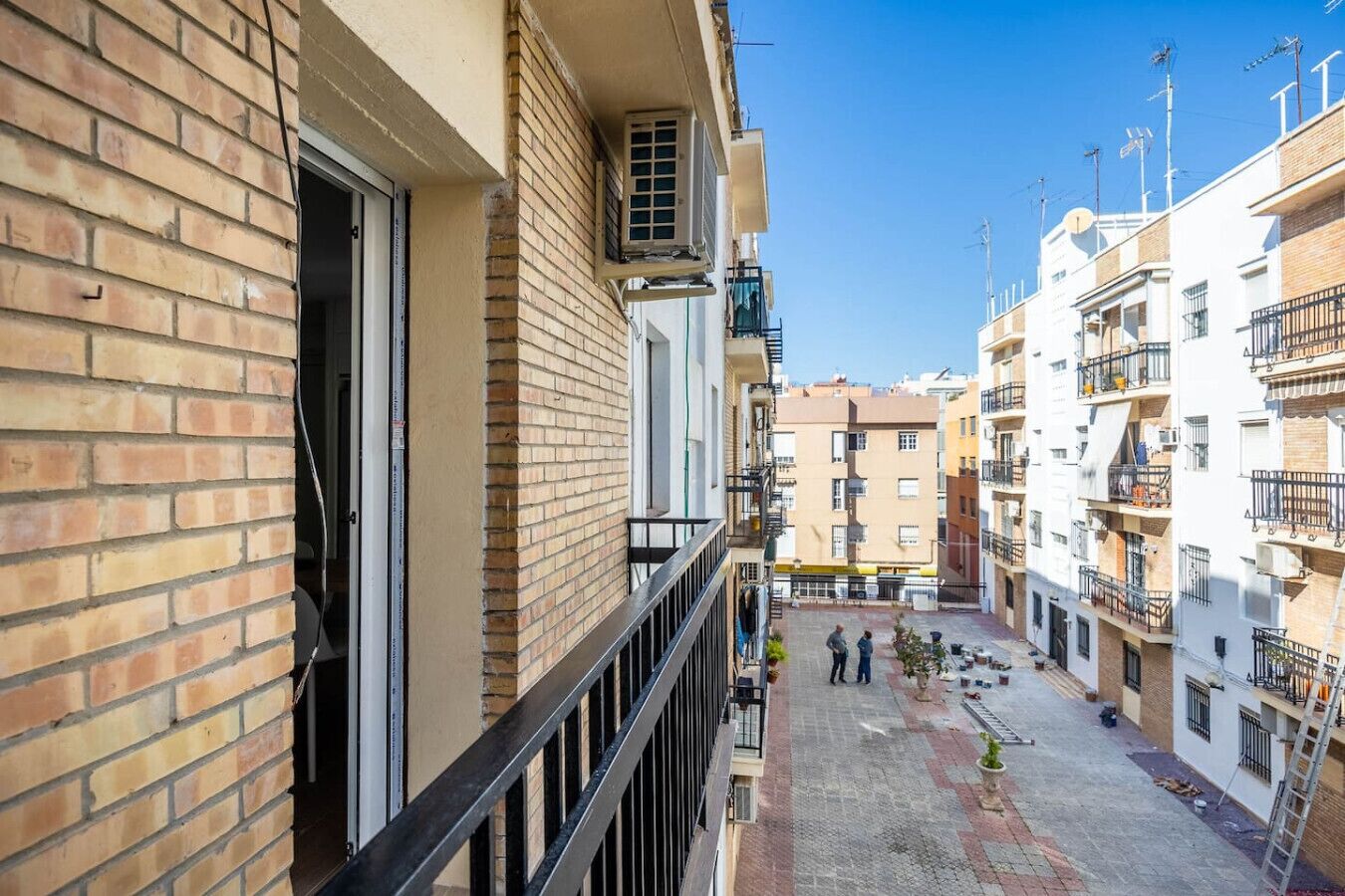 Une petite cour urbaine, avec un bâtiment en briques sur la gauche, un balcon doté d’une balustrade en métal et un ciel bleu dégagé.