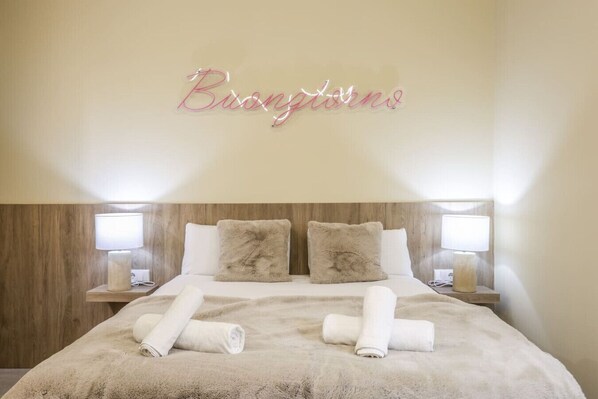 A hotel room with a bed, two fluffy pillows, two bedside lamps, and a neon sign above the bed.