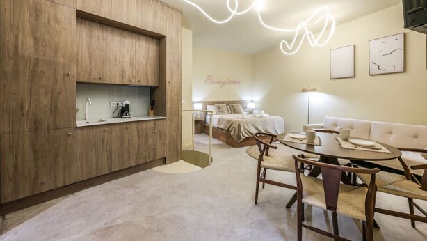 Une chambre d’hôtel moderne avec une armoire en bois, un coin repas comprenant une table ronde et des chaises, un lit avec une tête de lit blanche et un mur orné d’un néon.
