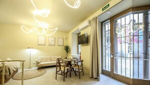 Une chambre d’hôtel comprenant un lit, un canapé, une table à manger avec des chaises, une lampe sur pied et une grande fenêtre avec des rideaux.