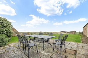 Un patio al aire libre con una mesa y sillas de metal, suelo de piedra y un muro de piedra.