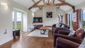 A spacious living room with a high ceiling, wooden beams, a chandelier, leather sofas, a flat-screen TV, and a large window offering a view of the outdoors.