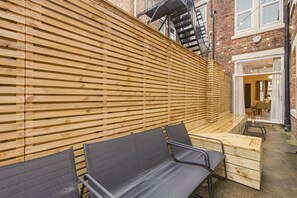 A wooden slatted wall, outdoor seating with metal chairs, and a brick building in the background.