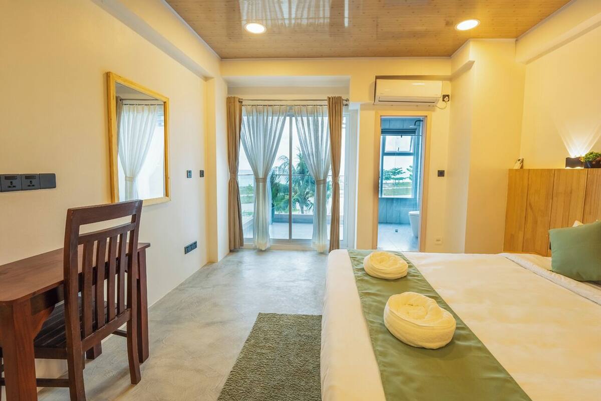 A hotel room with a bed, a desk, and a chair. There is a window with curtains, a door leading to an outdoor area, and a small plant on the bedside table.