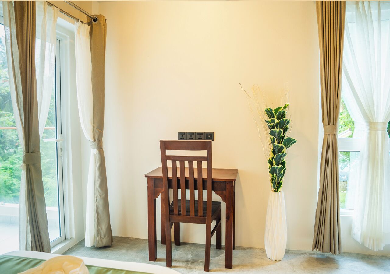 Una silla de madera con una pequeña mesa frente a una ventana con cortinas. Una planta en maceta está a la derecha de la silla.
