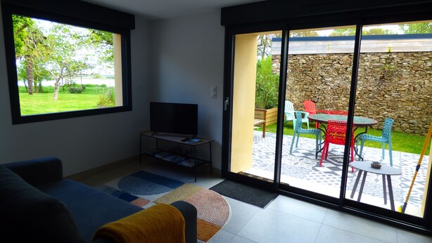 A living room with a TV, sofa, and a sliding glass door leading to an outdoor patio with chairs and a table. - Country house in Brière, 20 minutes from the beach. (Donges)