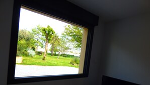 A room with a large window overlooking a grassy area with trees and a bench. - Country house in Brière, 20 minutes from the beach. (Donges)