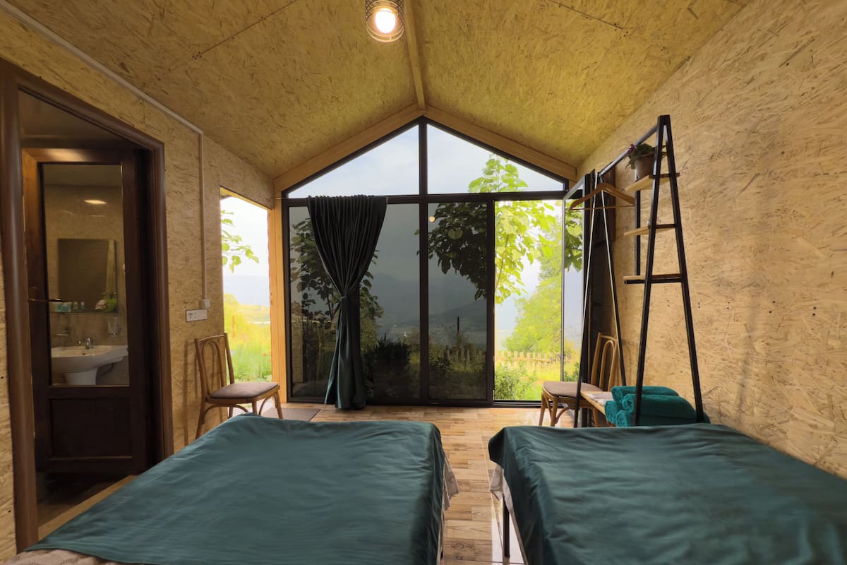 A room with two beds, a large window, a wooden ceiling, and a view of greenery outside.