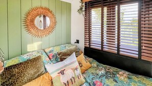 Un dormitorio con colcha estampada, varios almohadones con diseños variados, un espejo con forma de sol y una ventana con persianas de madera.