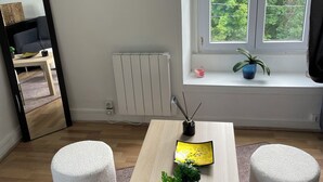 Une pièce avec une table en bois, deux poufs blancs, un radiateur, une fenêtre donnant sur un espace verdoyant, un tableau encadré au mur et une petite plante sur le rebord de la fenêtre.