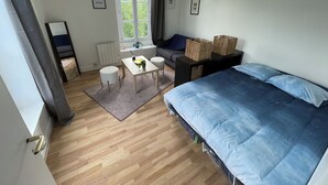 Une chambre à coucher avec un lit, une petite table et deux tabourets, une fenêtre avec des rideaux et un radiateur.