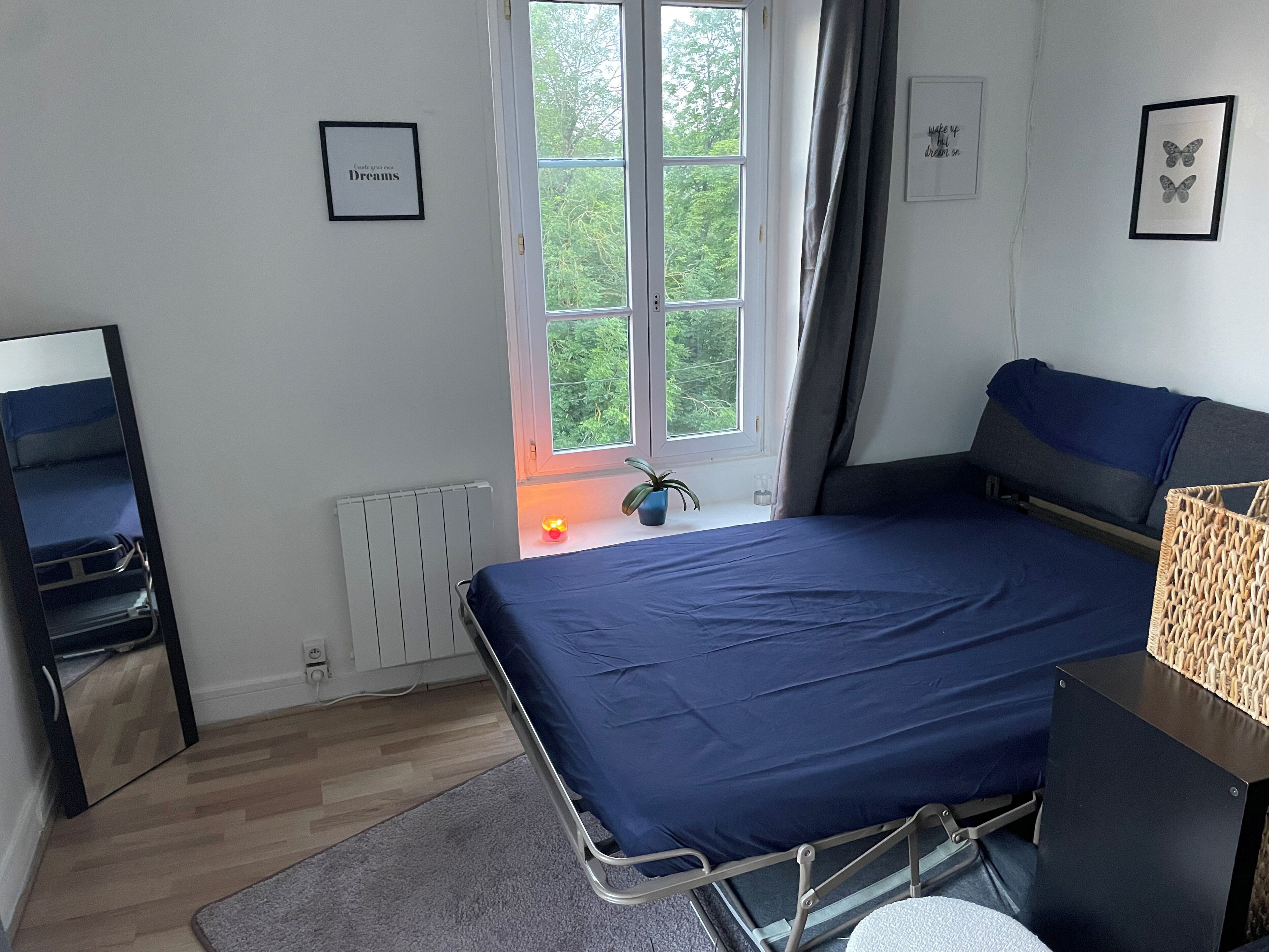 Een slaapkamer met een bed, nachtkastje, radiator, raam met gordijnen en een wand met ingelijste foto's.