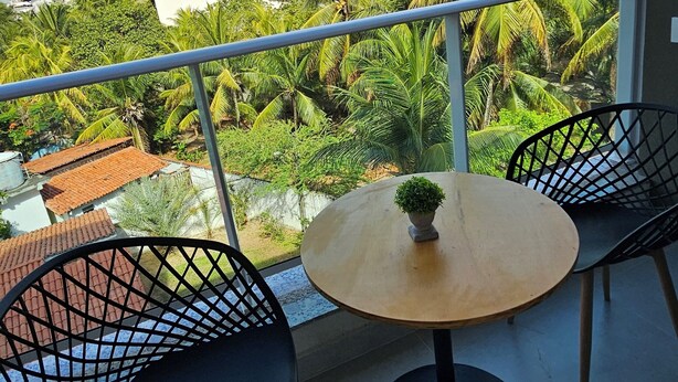 A balcony with two black chairs, a small round table, and a potted plant. The balcony overlooks a view of palm trees and buildings. - Conforto e localização Nixxus I (Ipojuca)