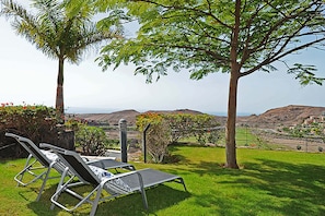 A grassy outdoor area with two lounge chairs, a palm tree, and a view of distant hills. - Salobre Morro Golf 9 (Maspalomas)