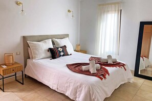 A neatly made bed with pillows, a bedside table with a lamp, and a window with curtains. - Salobre Morro Golf 9 (Maspalomas)