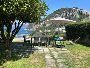 An outdoor seating area with a table and chairs, a large umbrella, and a view of the sea and mountains.