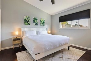 A bedroom with a bed, bedside tables, a ceiling fan, a window with a blind, and wall art.