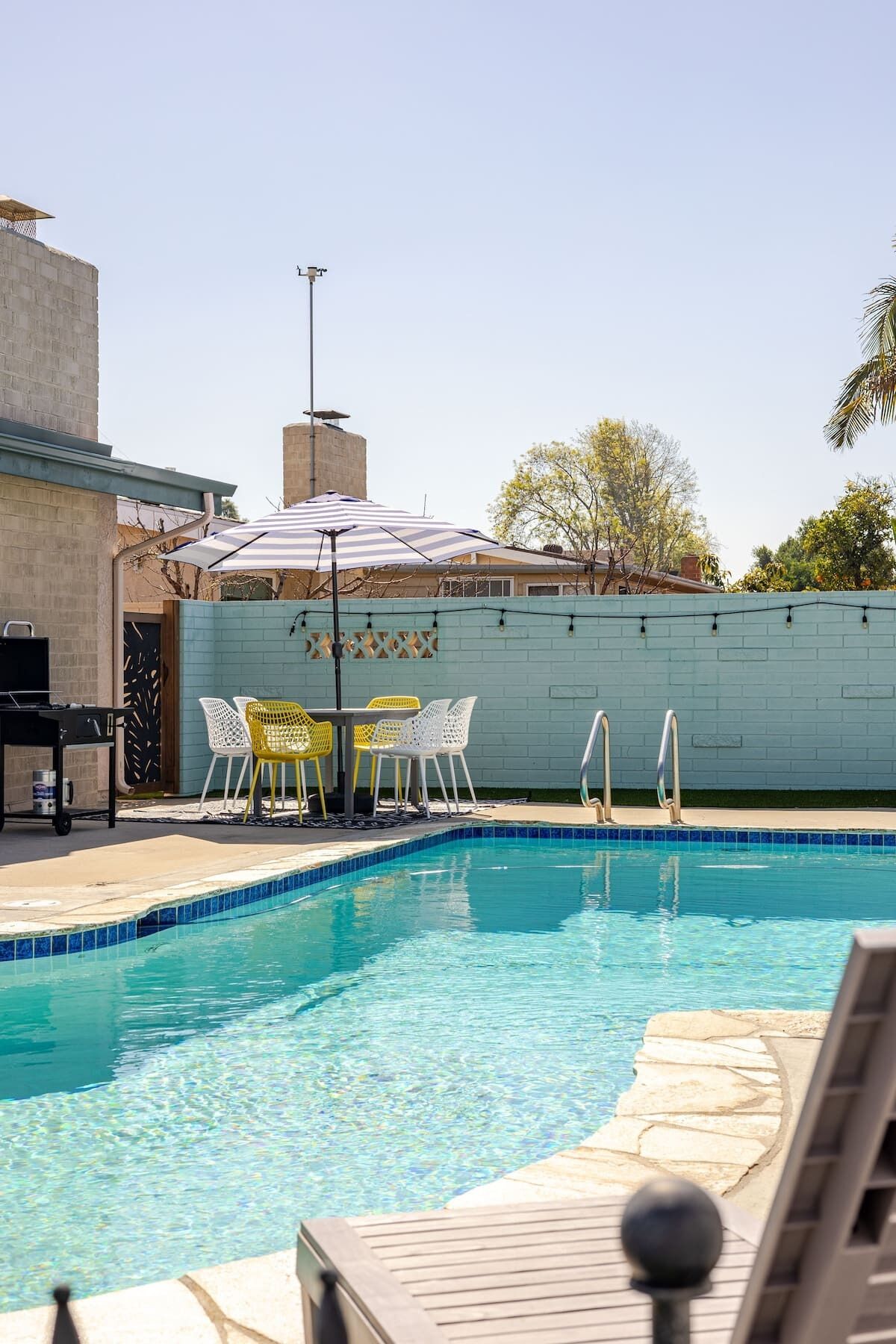 A swimming pool with a tiled edge, surrounded by a deck with lounge chairs. There is a table with chairs and a grill.