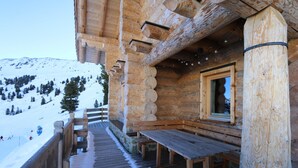 Eine Holzhütte mit einer Holzterrasse, Bänken und einem Fenster mit Blick auf einen verschneiten Berg.