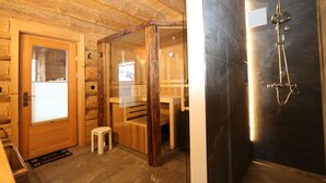 Ein Holzinnenraum mit Holztür, einem kleinen Hocker und einer Duschkabine mit dunkel gefliesten Wänden.