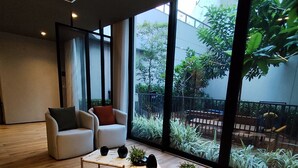 A modern interior with a wooden floor, a sofa, a coffee table, and large windows offering a view of greenery outside.