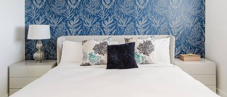 A bed with white bedding, a blue patterned wall, and a nightstand with a lamp and a book.