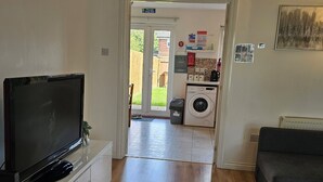 Ein Wohnzimmer mit Flachbildfernseher, einem grauen Sofa und Holzboden. Im Hintergrund ist eine Waschmaschine zu sehen.