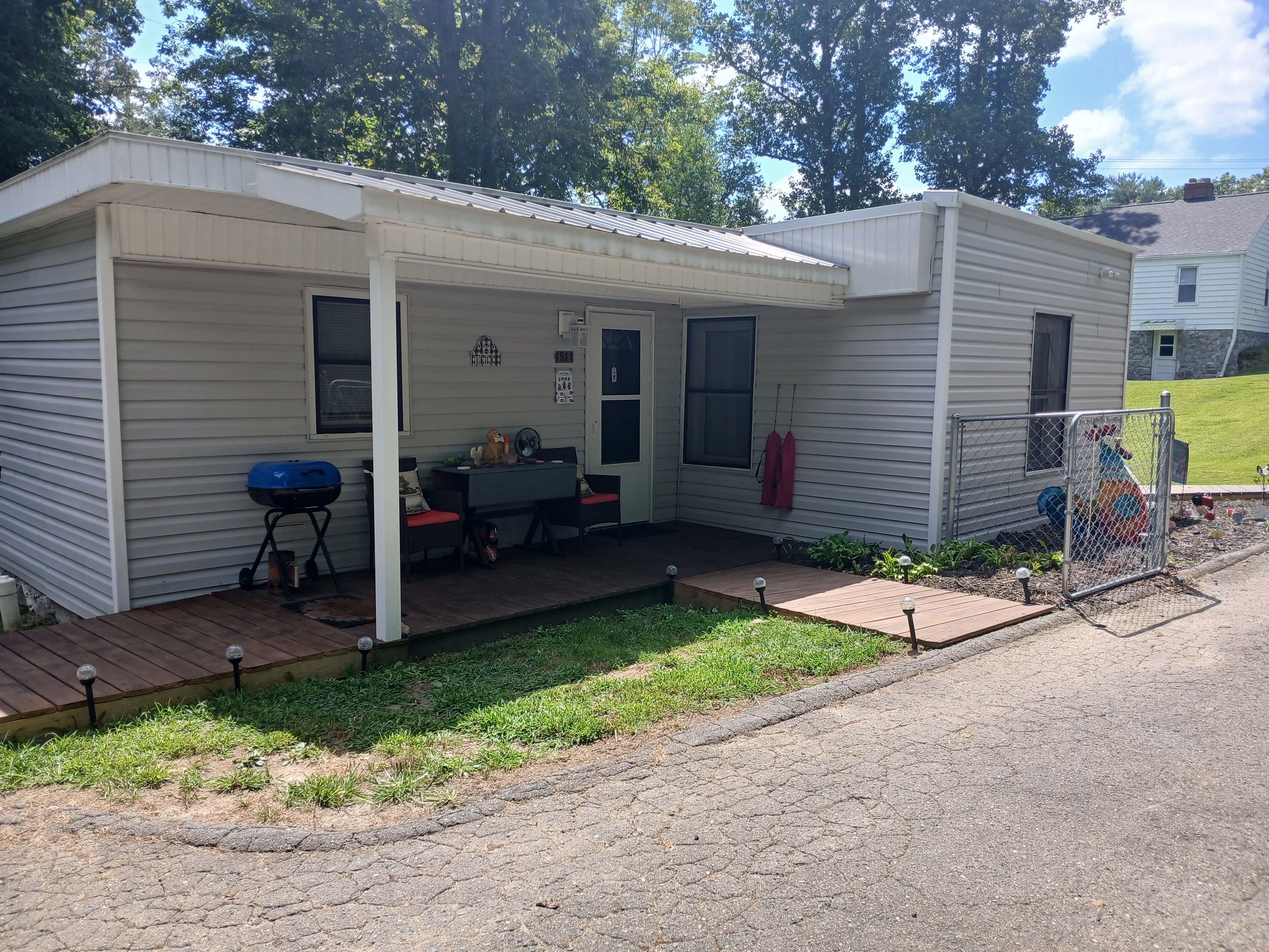 A small, single-story mobile home with a covered porch, a barbecue grill, and a fenced yard.