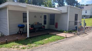 A small, single-story mobile home with a covered porch, a barbecue grill, and a fenced yard.