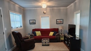 Un salon avec un canapé rouge, un fauteuil en cuir marron, une petite table d’appoint, une lampe, un meuble pour la télévision et un plancher en bois.