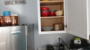 Une cuisine avec un réfrigérateur en acier inoxydable, un pichet en cuivre, une passoire jaune, des bols rouges et beiges, et un panneau sur lequel on peut lire : « VIVEZ. AIMEZ. AYEZ. »