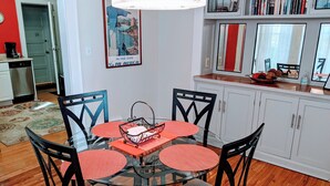 Un coin repas avec une table en verre entourée de chaises, une cuisine en arrière-plan et une bibliothèque remplie de livres.