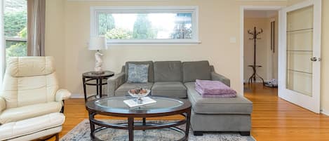 A living room with a leather chair, a grey sofa, a glass coffee table, and a window with a view of greenery.
