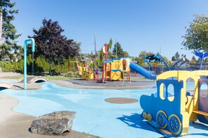 A playground with a blue and yellow train-themed play structure, a water play area, and a large rock in the foreground. - The Forrester Vista (Oak Bay North)