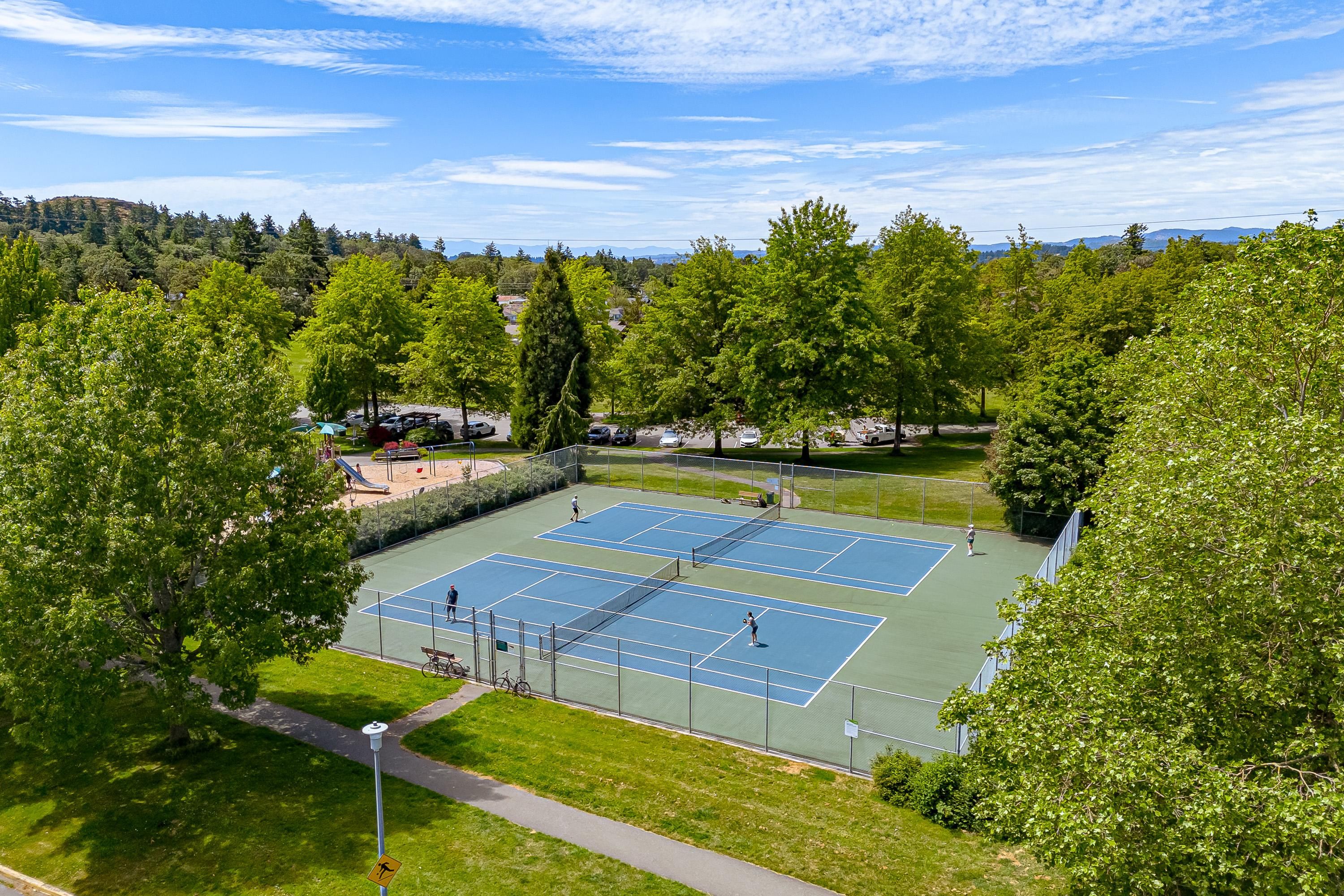 A tennis court surrounded by trees and a parking area.