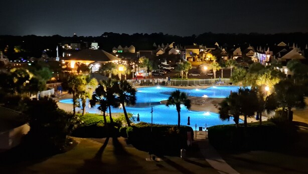 Vista noturna de um resort com piscina, palmeiras e edifícios iluminados.