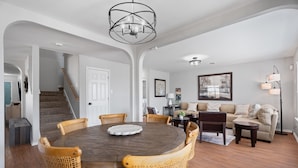 A dining area with a round wooden table and wicker chairs, a staircase, a living room with a sofa and lamp, and a hallway with a door. - Spacious 5BR / Perfect for Families & Groups (Universal City)