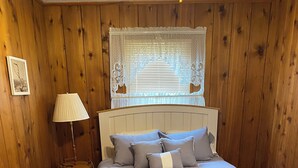 Ein Schlafzimmer mit Holzboden, einem Bett mit grauer Bettwäsche, einer Nachttischlampe und einem Fenster mit Spitzenvorhängen.