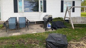 Ein weißes Haus mit schwarzen Fensterläden, einem Grill und zwei Stühlen im Garten.