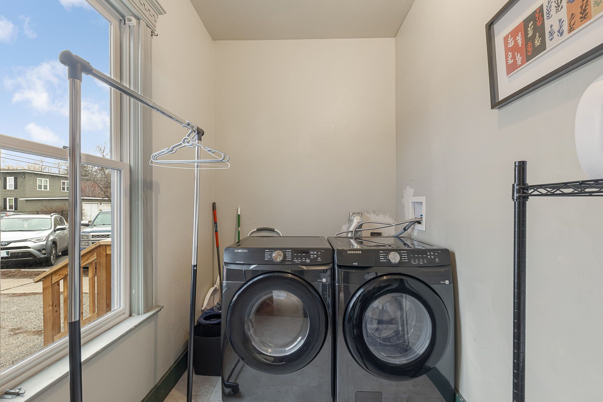 A laundry room with two Samsung washing machines, a drying rack, and a window with a view of parked cars and houses outside.