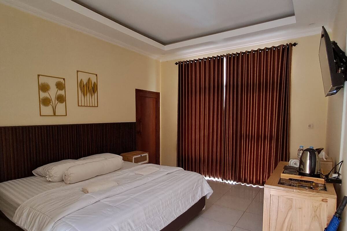 A hotel room with a large bed, a wooden desk, a television, and a window with curtains.