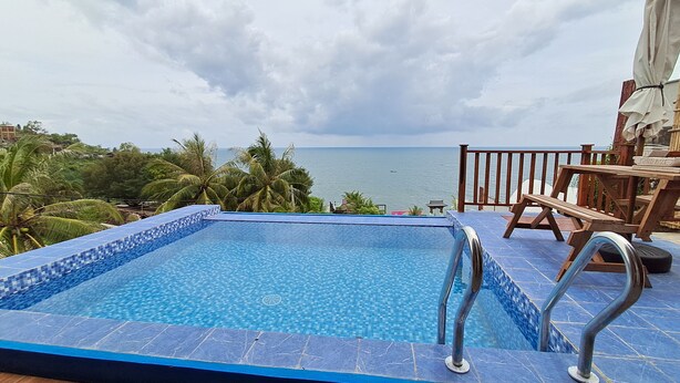 A pool with a view of the sea, surrounded by palm trees and a wooden deck with a table and chairs. - Teras Kaca (Panggang)