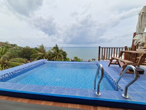 A pool with a view of the sea, surrounded by palm trees and a wooden deck with a table and chairs.