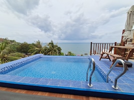 Piscina com vista para o mar, rodeada por palmeiras e um deck de madeira com mesa e cadeiras.