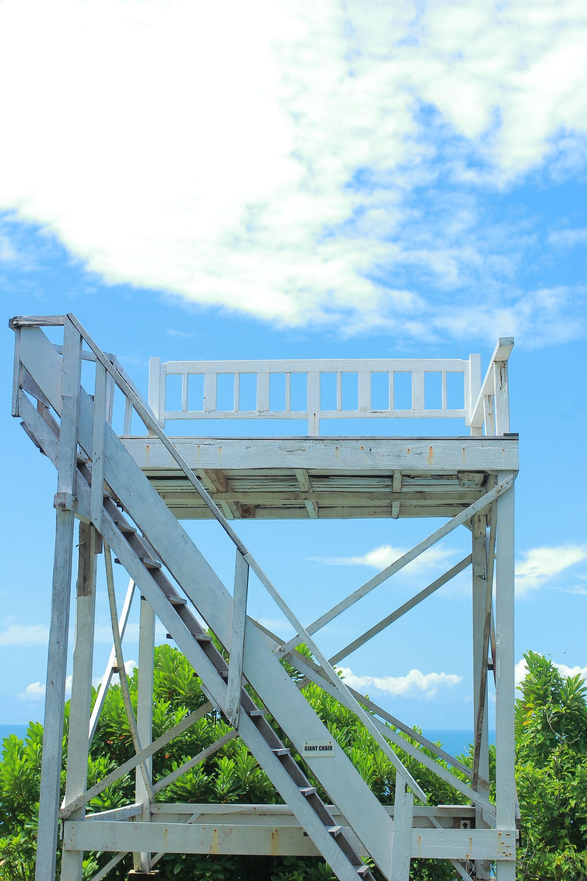 A white metal structure with a bench, surrounded by greenery and a clear sky.