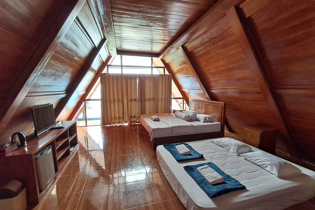 A room with a sloped ceiling, wooden walls, a bed with pillows, a TV, and a small table with a kettle.