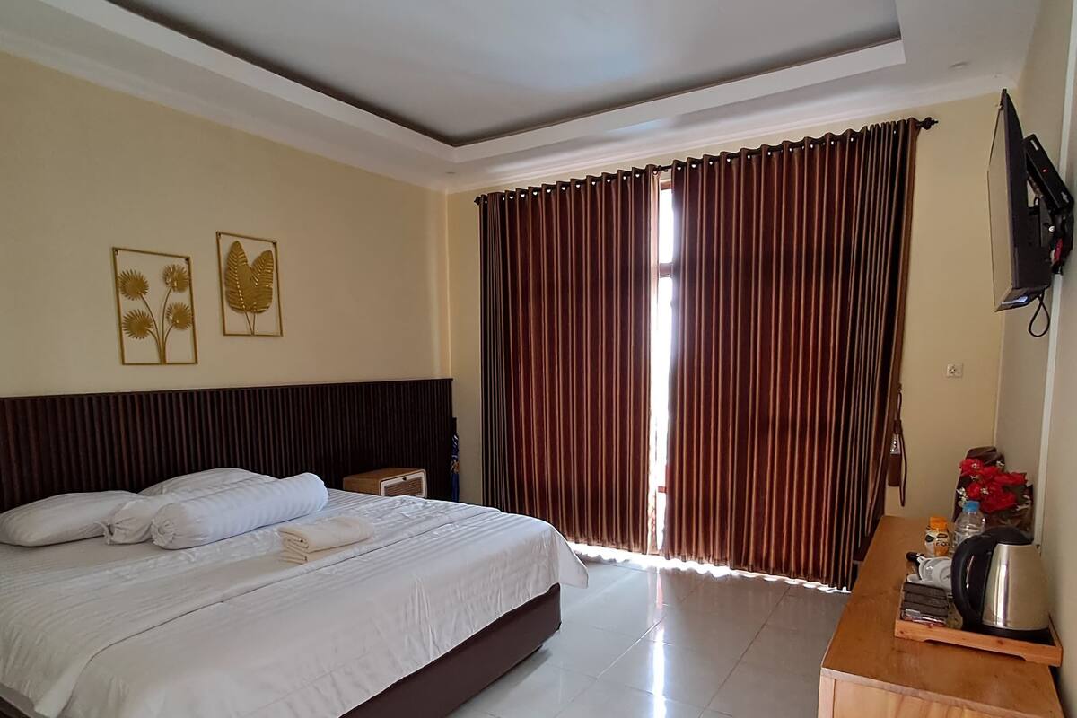 A hotel room with a large bed, a wooden nightstand, a desk with a kettle, and a TV on the wall.